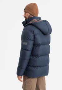 Giubbotto trapuntato blu navy con cappuccio, caratterizzato da sezioni trapuntate orizzontali, un berretto in maglia marrone texturizzato e una fodera interna a quadri.