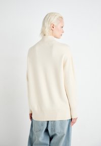 Cremfarbener Pullover mit hohem Kragen, gerippten Bündchen und Saum. Über lockeren hellblauen Jeans getragen. Weiches, strukturiertes Maschenmaterial.