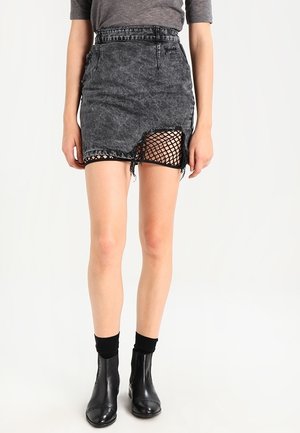 Donna che indossa una minigonna nera effetto acid wash con un dettaglio strappato che rivela un tessuto a rete nero sotto, abbinata a stivaletti e calzini neri.