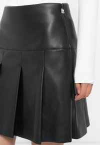 Gonna di pelle nera a pieghe indossata con un top bianco a maniche lunghe, con cerniera laterale in vista e una mano che poggia vicino all'orlo.