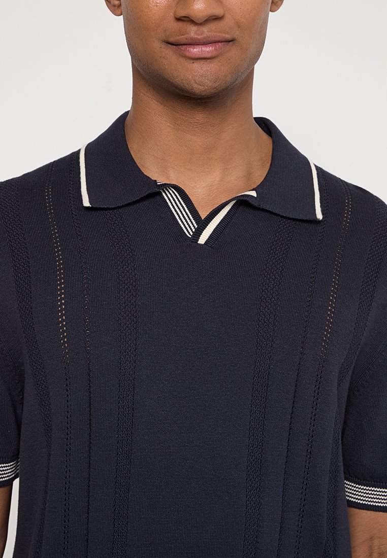 Homme portant un polo en tricot bleu marine avec des rayures blanches sur le col et les bords des manches, sur fond clair uni.