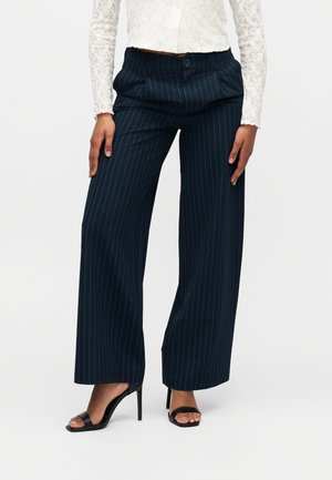 Γυναίκα που φοράει navy μπλε παντελόνι με ρίγες και φαρδιά πόδια, συνοδευόμενο από μαύρα πέδιλα με ψηλό τακούνι, και λευκό μακρυμάνικο τοπ από δαντέλα.