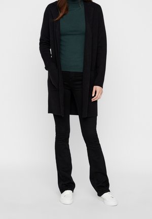 Schwarzer, langer Cardigan mit offener Vorderseite, zwei Seitentaschen, getragen über einem dunkelgrünen Rollkragenpullover, kombiniert mit schwarzen, ausgestellten Jeans und weißen Turnschuhen.