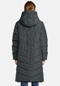 Femme portant un long manteau d'hiver matelassé noir avec une capuche, se tenant de dos contre un fond clair.