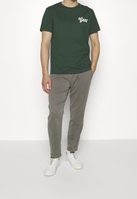 Zelené bavlněné tričko s bílým logem "Gant", kombinované se světle šedými kalhotami a bílými teniskami. Jednoduchý, neformální outfit bez vzorů.