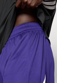 Μωβ αθλητικές σορτς από δίχτυ με ελαστική μέση, συνδυασμένα με μαύρο πουκάμισο που εμφανίζει διακριτή υφή και πλαϊνές ρίγες.