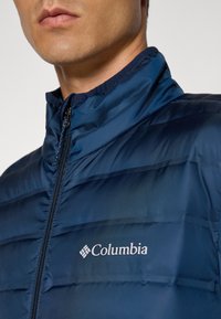 Tamsiai mėlyna pūkinė striukė su aukštu apykakle, užtrauktuku ir horizontaliais siūlėmis. Columbia logotipas matomas ant krūtinės.