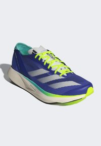 Zapatilla deportiva azul con un upper de malla texturizada, que presenta franjas reflectantes plateadas, acentos en verde neón y una suela ligera.