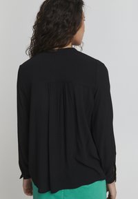Blusa nera a maniche lunghe in tessuto testurizzato, con dettaglio sul retro a yoke e polsini con bottoni, abbinata a una gonna verde, che mostra un design a strati.