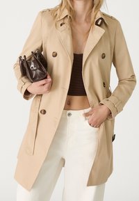 Trench beige con botones de doble botonadura, hombreras y bolsillos laterales. El modelo sostiene un pequeño bolso marrón. Jeans blancos visibles.