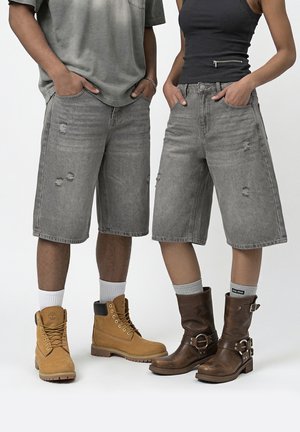 Deux personnes portant des shorts en jean gris usés jusqu'aux genoux, debout les mains dans les poches, bottes de travail couleur beige et bottes marron à boucle.