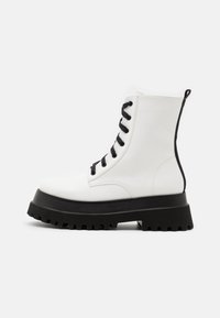 Hvide snørebåndsko til anklerne med sort chunky sål, lavet af glat syntetisk materiale. Funktionen inkluderer sorte øjer og en løkkesnor bagpå.