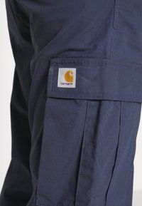 Námořnické modré cargo kalhoty vyrobené z texturované látky, s nášivkou loga Carhartt v oranžové a bílé barvě na boční kapse.