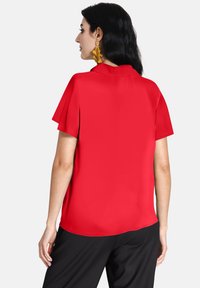 Blusa rossa a maniche corte con colletto classico, texture liscia e vestibilità ampia, abbinata a pantaloni neri e grandi orecchini gialli.