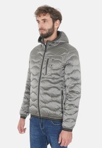 Giacca imbottita grigia trapuntata con cappuccio, chiusura a zip frontale e tasca, polsini ed orlo elasticizzati. Materiale morbido e lucido con motivo testurizzato.