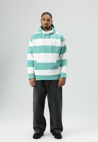 Turquoise-wit gestreepte hoodie met een voorzak, capuchon met trekkoord en een losse pasvorm. Draagt bij donkere wijde jeans en zwarte sneakers.