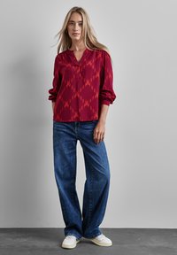 Blusa a motivi rossi e arancioni con scollatura a V e maniche lunghe, abbinata a jeans blu larghi e scarpe da ginnastica bianche, su uno sfondo neutro.