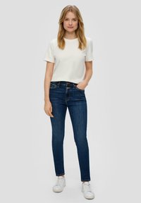 T-shirt blanc à manches courtes associé à un jean skinny bleu foncé et des baskets blanches, mettant en avant un design décontracté et ajusté.