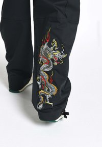 Zwarte broek met gedetailleerde draakborduursel in levendige kleuren, met accenten van rood, geel en grijs aan de zijkant. Zachte textuur.