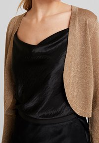 Goldfarbenes Strick-Cardigan aus Metallicgarn mit kurzem, offenem Schnitt. Getragen über einem glatten schwarzen Satinoberteil mit drapiertem Ausschnitt.