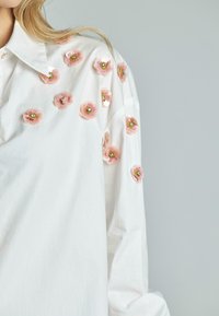 Camicia bianca con applicazioni floreali rosa e accenti dorati sulle spalle. Il tessuto appare liscio e leggero.