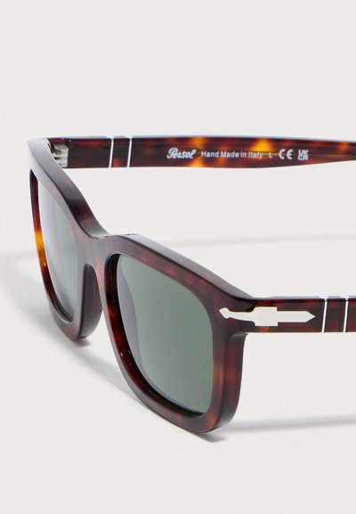 Τετράγωνα γυαλιά ηλίου σε σχέδιο χελωνακίου με σκουρό πράσινους φακούς, λευκή λεπτομέρεια βέλους στα πλαϊνά και την επιγραφή "Persol Hand Made in Italy" στο εσωτερικό του βραχίονα.