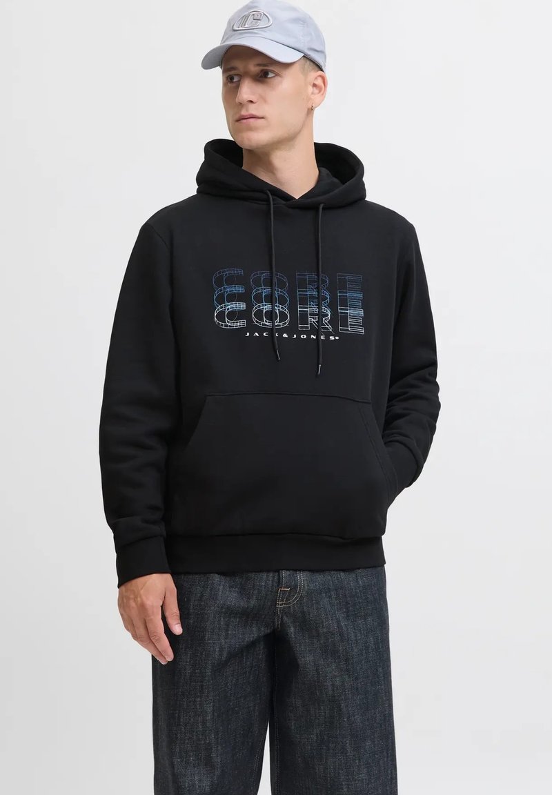 Uomo in felpa nera con testo azzurro "CORE", jeans scuri e cappello grigio chiaro, che guarda di lato con una mano in tasca.