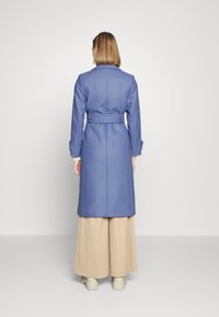 Femme aux cheveux mi-longs portant un manteau bleu ceinturé, un pantalon beige à large jambe et des chaussures à plateforme crème, tournée de dos par rapport à l'objectif.