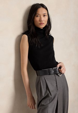 Vrouw met lang zwart haar, die een zwarte mouwloze coltrui en hoog getailleerde grijze geplooide broek met brede zwarte riem draagt, staat tegen een beige muur.