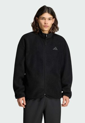 Ung person med langt mørkt hår iført en sort fleecejakke med lynlås og et diskret Adidas-logo på brystet samt sorte bukser.