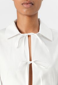 White blouse featuring a collar and tie closures at the neckline. The fabric appears smooth with a crisp texture. Cut-out detail at the front.