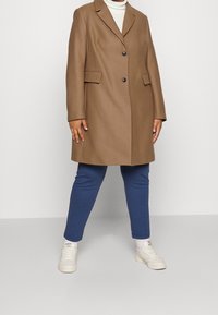Brown wool blend overcoat with a notch lapel, two front pockets, and a double button closure, paired with blue slim-fit pants and white sneakers.