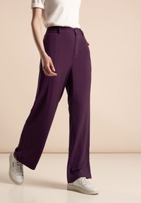 Pantalons larges violets en tissu lisse, avec une taille élastique et une fermeture à bouton à l'avant, assortis à des baskets blanches.