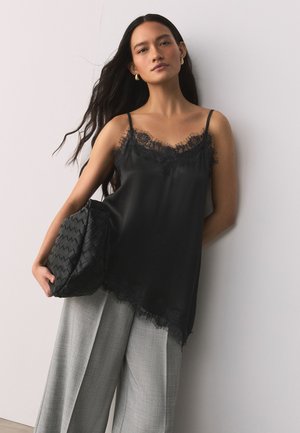 Femeie cu păr lung și închis la culoare, purtând un maiou negru cu dantelă, pantaloni largi gri, ținând o geantă plic neagră texturată în fața unui perete deschis.