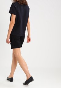 Camiseta de algodón negra de manga corta, combinada con pantalones cortos negros. El modelo lleva alpargatas negras; se ve desde la espalda, dando la espalda.