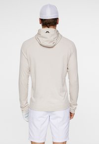 Beige långärmad hoodie med logotyp på nacken, mjukt tyg, fast huva och snedställda raglanärmar, parat med vita shorts.