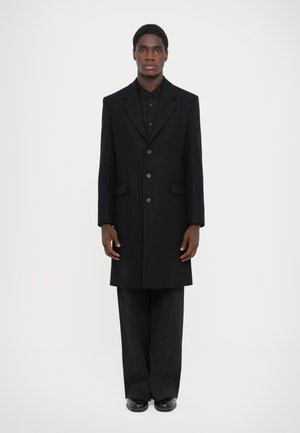 Lång svart ullkappa med notch-lapel, framfickor och knäppning, bärs över en svart mönstrad skjorta och svarta byxor.