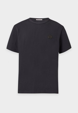 Ung mand stående med hænderne i lommerne iført en sort T-shirt med korte ærmer, sorte bukser og sorte sko mod en hvid baggrund.