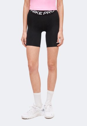 Mujer con pantalones cortos Nike Pro negros, una camiseta rosa, calcetines blancos hasta el tobillo y zapatillas deportivas blancas, de pie frente a un fondo claro y sencillo.