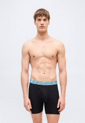 Noor mees, kellel on lühikesed pruunid juuksed, seljas mustad bokserid helesinise vööosaga, seisab ühevärvilise helehall tausta ees.