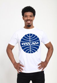 T-shirt bianca in cotone con una grande grafica circolare blu con linee e il testo "PAN AM" in un carattere prominente. Il modello sorride, con le mani sui fianchi.