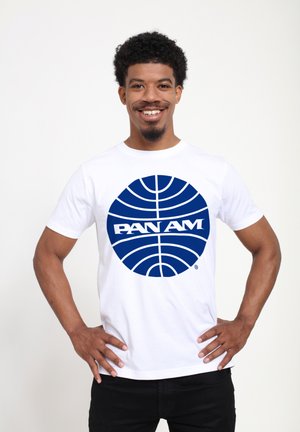 Bílá bavlněná trička s velkým modrým kruhovým grafickým motivem s linkami a nápisem "PAN AM" ve výrazném písmu. Modelka se usmívá a má ruce na bocích.