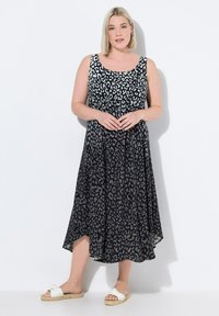Schwarzes ärmelloses Kleid mit einem grauen Leopardenmuster, A-Linie-Form, fließender Stoff, knielanger Saum, kombiniert mit weißen Sandalen mit einer Schleifenverzierung.