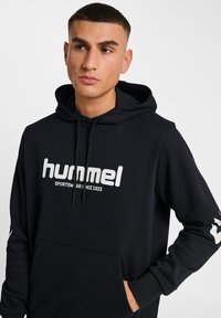 Svart huvtröja med en stor vit "hummel"-logotyp. Har en framficka, dragsnoddar och vita detaljer på ärmarna.