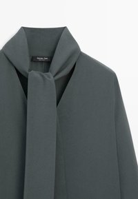 Blusa grigia antracite con colletto drappeggiato e dettaglio con nodo in tessuto. Texture liscia e design minimalista, caratterizzata da un'etichetta di marca discreta.