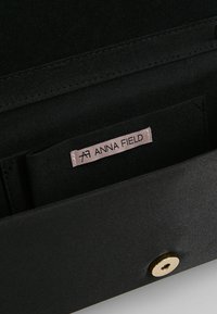 Svart tygplånbok med en jämn textur, med en guldknappstängning och en synlig etikett som läser "ANNA FIELD." Inuti har den en enkel design.