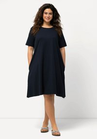 Námořnická modrá šatová košile vyrobená z lehkého materiálu, s krátkými rukávy, volným střihem a bočními kapsami. Nosená s šedými sandály.