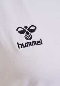 White cotton shirt featuring a black embroidered logo of a stylized bee with the text "hummel" underneath. Smooth texture, classic fit.