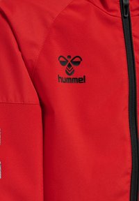 Giacca rossa realizzata in tessuto liscio. Presenta una cerniera nera e il logo Hummel in nero nella parte superiore sinistra.