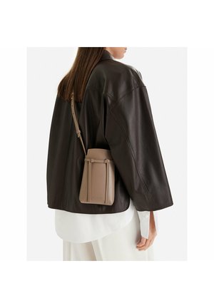 Kobieta w ciemnobrązowej, oversize'owej skórzanej kurtce, nosząca beżową torbę na ramię przewieszoną przez białą koszulę i spodnie, pokazana z tyłu i z boku.